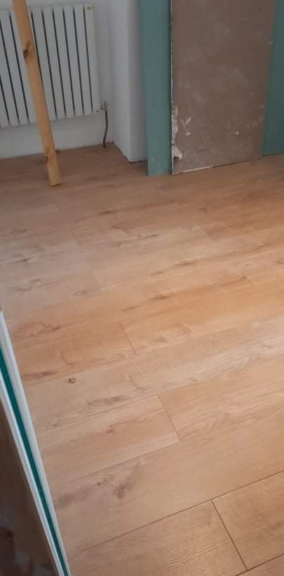 Jasne panele podłogowe ułożone w nowym pomieszczeniu, widoczny fragment grzejnika i przygotowana ściana do dalszych prac wykończeniowych.