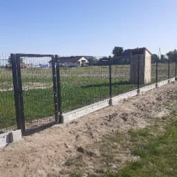 Nowoczesne ogrodzenie panelowe z bramką, osadzone na betonowej podmurówce z elementami dekoracyjnymi, widok na teren z domem w tle.