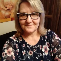 Portret kobiety w okularach z dwukolorową oprawką, w kwiecistej bluzce na tle ściany z drewnianą boazerią i obrazem róży.