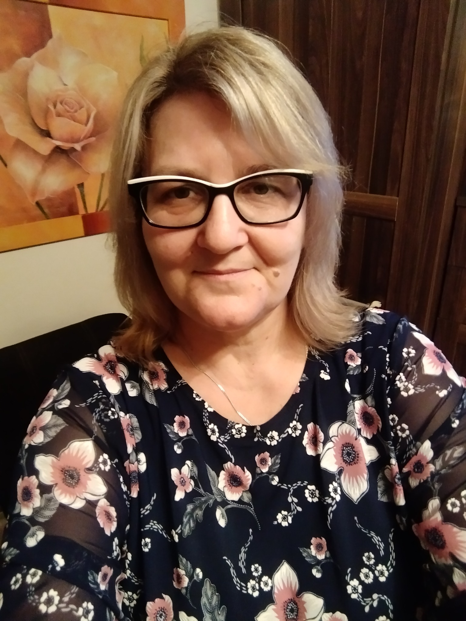 Portret kobiety w okularach z dwukolorową oprawką, w kwiecistej bluzce na tle ściany z drewnianą boazerią i obrazem róży.