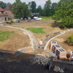 Budowa ścieżki z kostki brukowej na terenie posesji z widocznym budynkiem w tle, materiałami budowlanymi i pracownikiem z wiadrami.