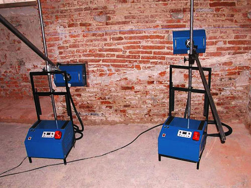 Dwa niebieskie osuszacze kondensacyjne na tle ceglanej ściany z widocznymi śladami wilgoci.