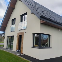 SEBASTIAN MILEWSKI - Elewacja domu jednorodzinnego z grafitową dachówką i stolarką okienną. Widoczne szklane balustrady balkonowe i wykończenie wokół drzwi wejściowych imitujące drewno.