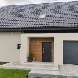 SEBASTIAN MILEWSKI - Nowoczesny dom z grafitową dachówką, jasną elewacją i drewnianym akcentem przy wejściu. Uwagę zwraca brama garażowa w kolorze antracytu.