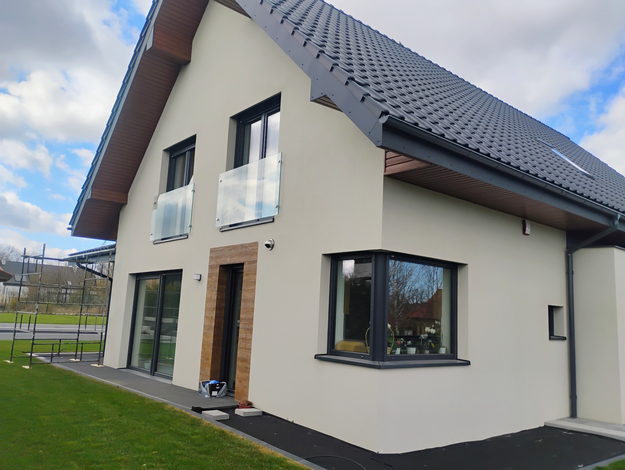 Elewacja domu jednorodzinnego z grafitową dachówką i stolarką okienną. Widoczne szklane balustrady balkonowe i wykończenie wokół drzwi wejściowych imitujące drewno.