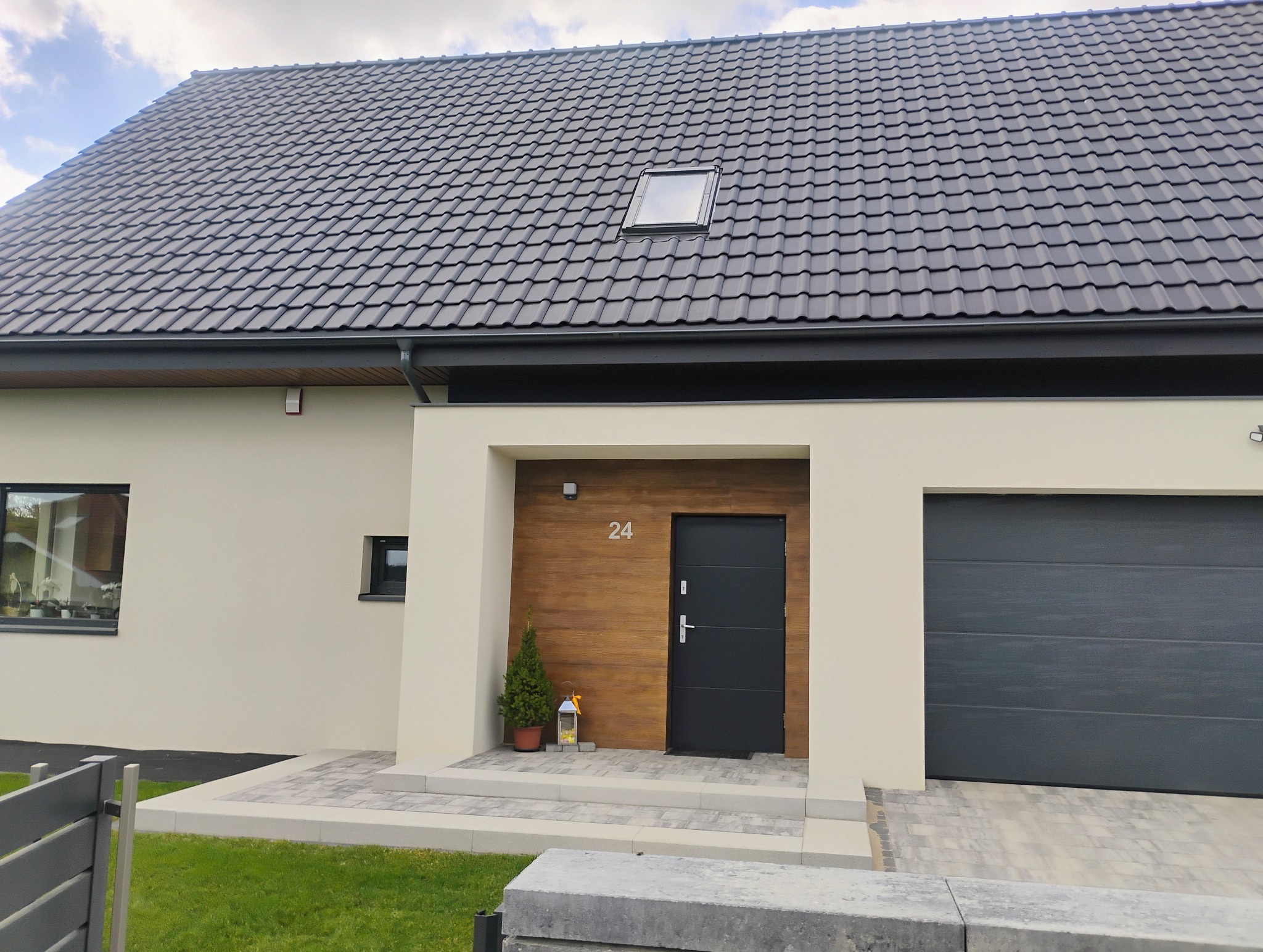 Nowoczesny dom z grafitową dachówką, jasną elewacją i drewnianym akcentem przy wejściu. Uwagę zwraca brama garażowa w kolorze antracytu.