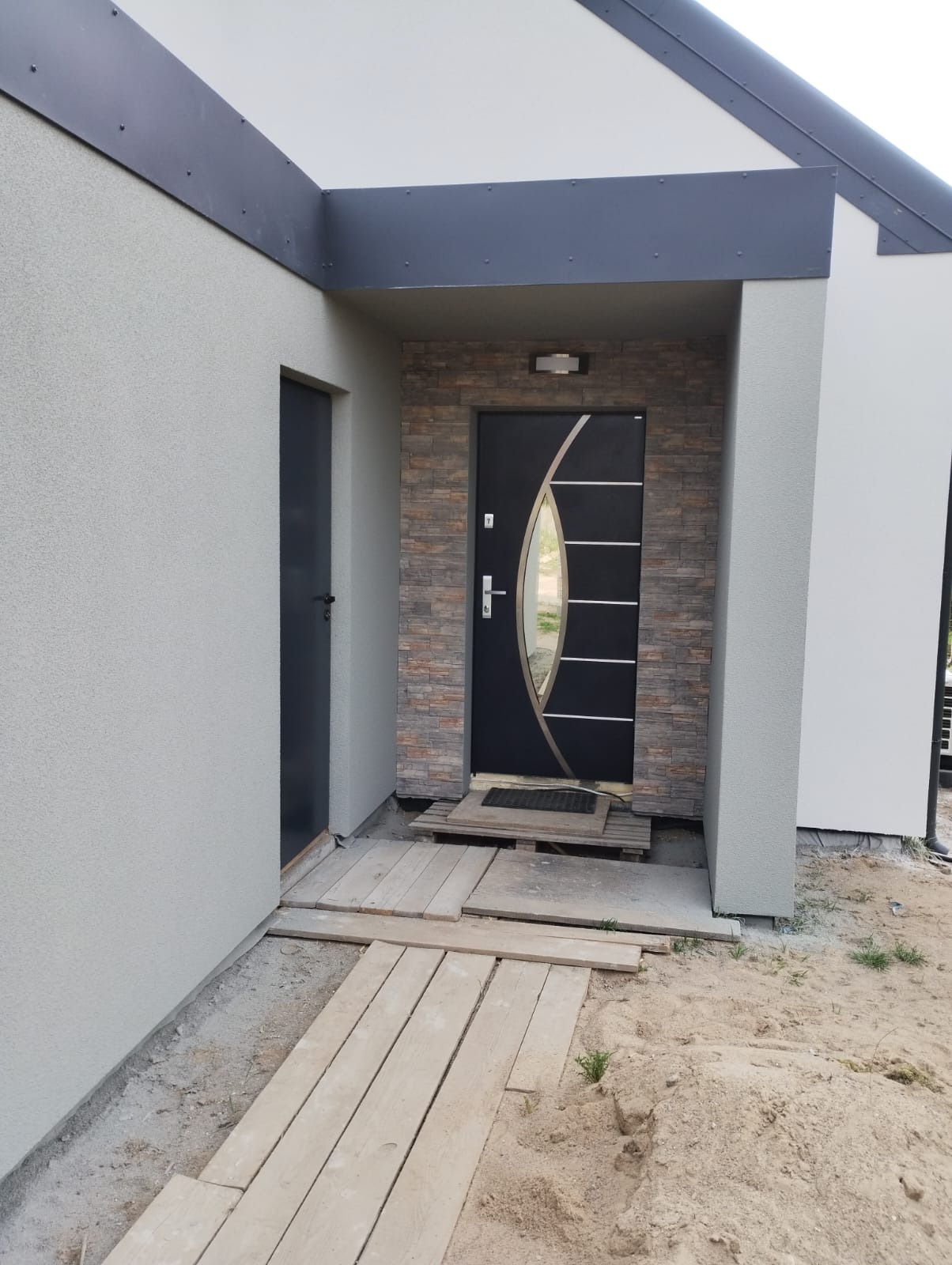 Nowoczesne wejście do domu z czarnymi drzwiami z szybą i okładziną z kamienia elewacyjnego. Widoczny tymczasowy chodnik z desek i fragment elewacji.