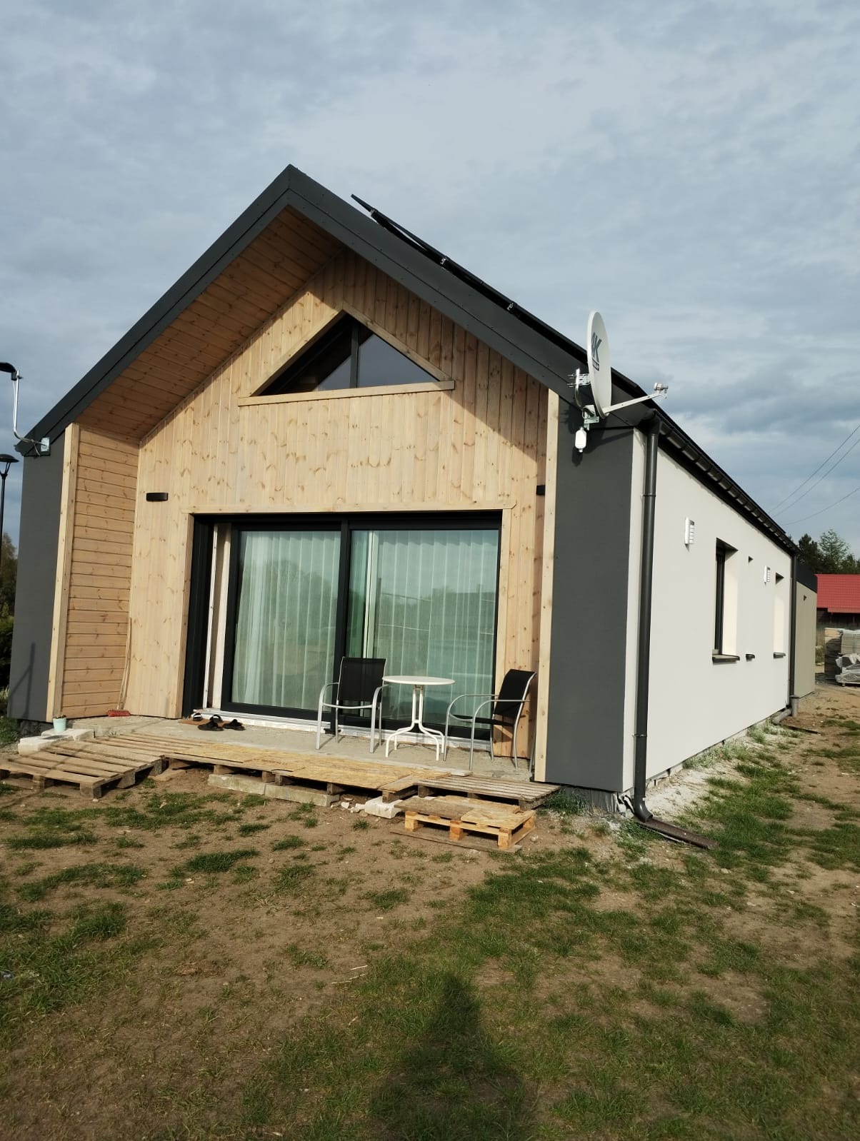 Nowoczesny dom z drewnianą elewacją i dużymi oknami, taras z palet, antena satelitarna na dachu. Kontrastowe kolory elewacji, minimalistyczny design.