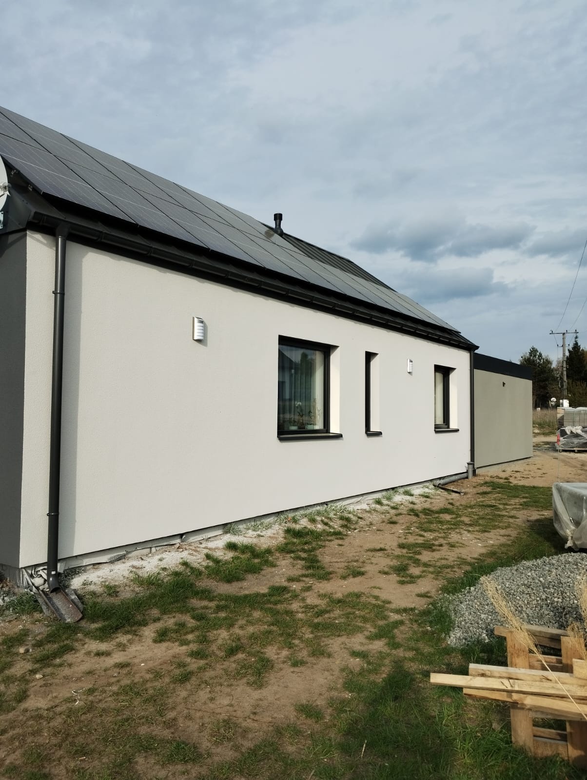 Elewacja domu jednorodzinnego z panelami solarnymi, widok z boku, z czarnymi ramami okiennymi i orynnowaniem. Na ziemi widoczne ślady po budowie i palety.