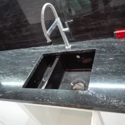 Czarny, polerowany blat z kamienia naturalnego z wbudowanym zlewozmywakiem dwukomorowym i nowoczesną baterią kuchenną ze sprężyną. Na blacie poziomica. Widoczne białe szafki pod blatem.