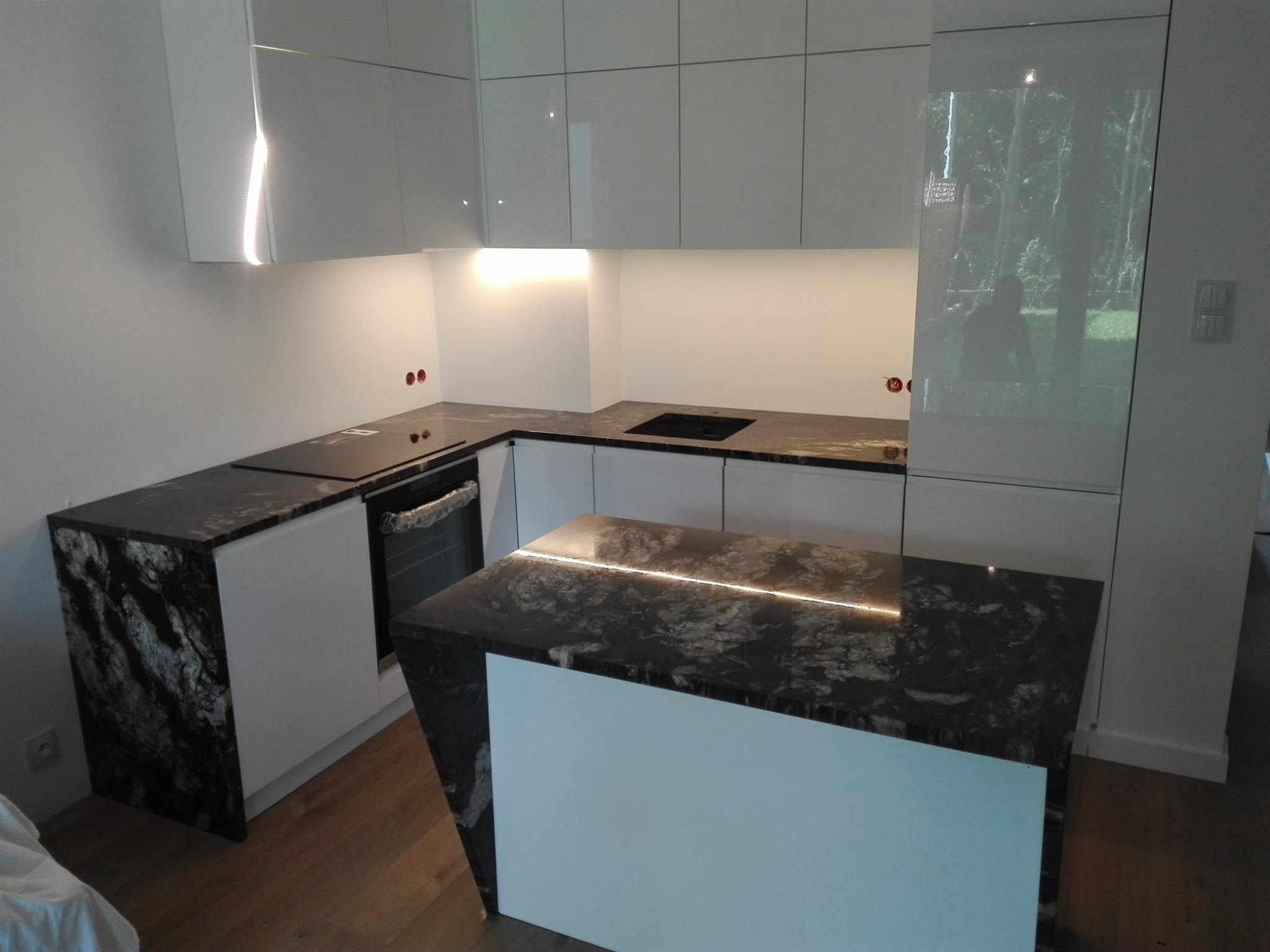 Nowoczesna kuchnia z czarnym blatem o wyraźnym wzorze żyłkowania, białymi szafkami i podświetleniem LED pod szafkami wiszącymi oraz na wyspie kuchennej.