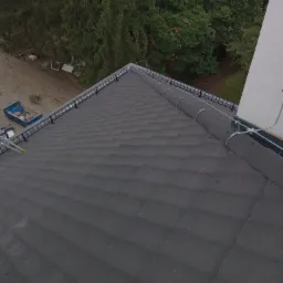 Fragment dachu pokrytego ciemną dachówką karpiówką, z widocznym systemem odgromowym i koszem dachowym, na tle zieleni drzew i fragmentu białej ściany budynku.