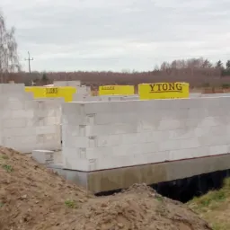 Wznoszenie ścian parteru z bloczków Ytong na fundamencie, widoczny stos materiałów budowlanych w żółtym opakowaniu, ziemia i drzewa w tle.