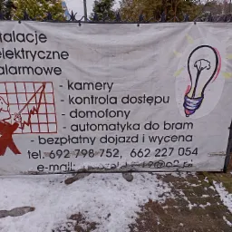 Biała, lekko zabrudzona banerowa reklama usług elektrycznych i alarmowych, zamocowana na metalowym ogrodzeniu, z grafiką żarówki i schematyczną postacią elektryka, widoczna spod śniegu.