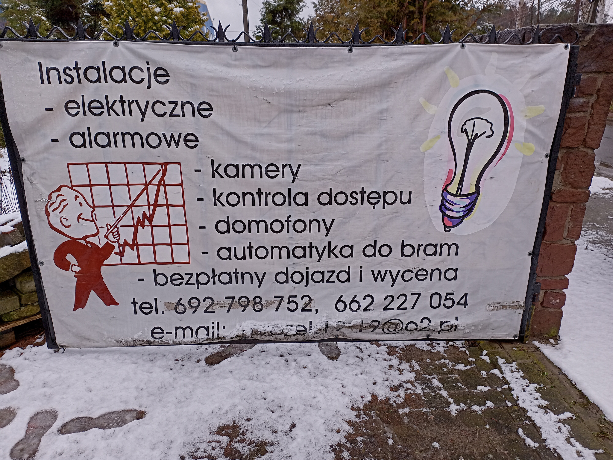 Biała, lekko zabrudzona banerowa reklama usług elektrycznych i alarmowych, zamocowana na metalowym ogrodzeniu, z grafiką żarówki i schematyczną postacią elektryka, widoczna spod śniegu.