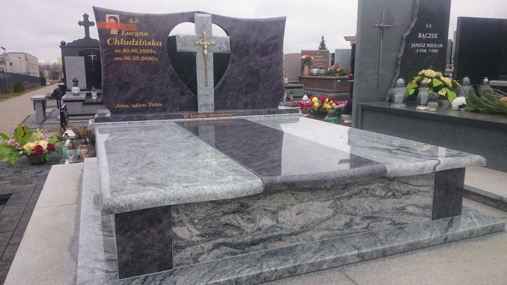 Granitowy pomnik z krzyżem w kształcie serca, z wygrawerowanym napisem i datami, otoczony kwiatami i lampionami na cmentarzu.