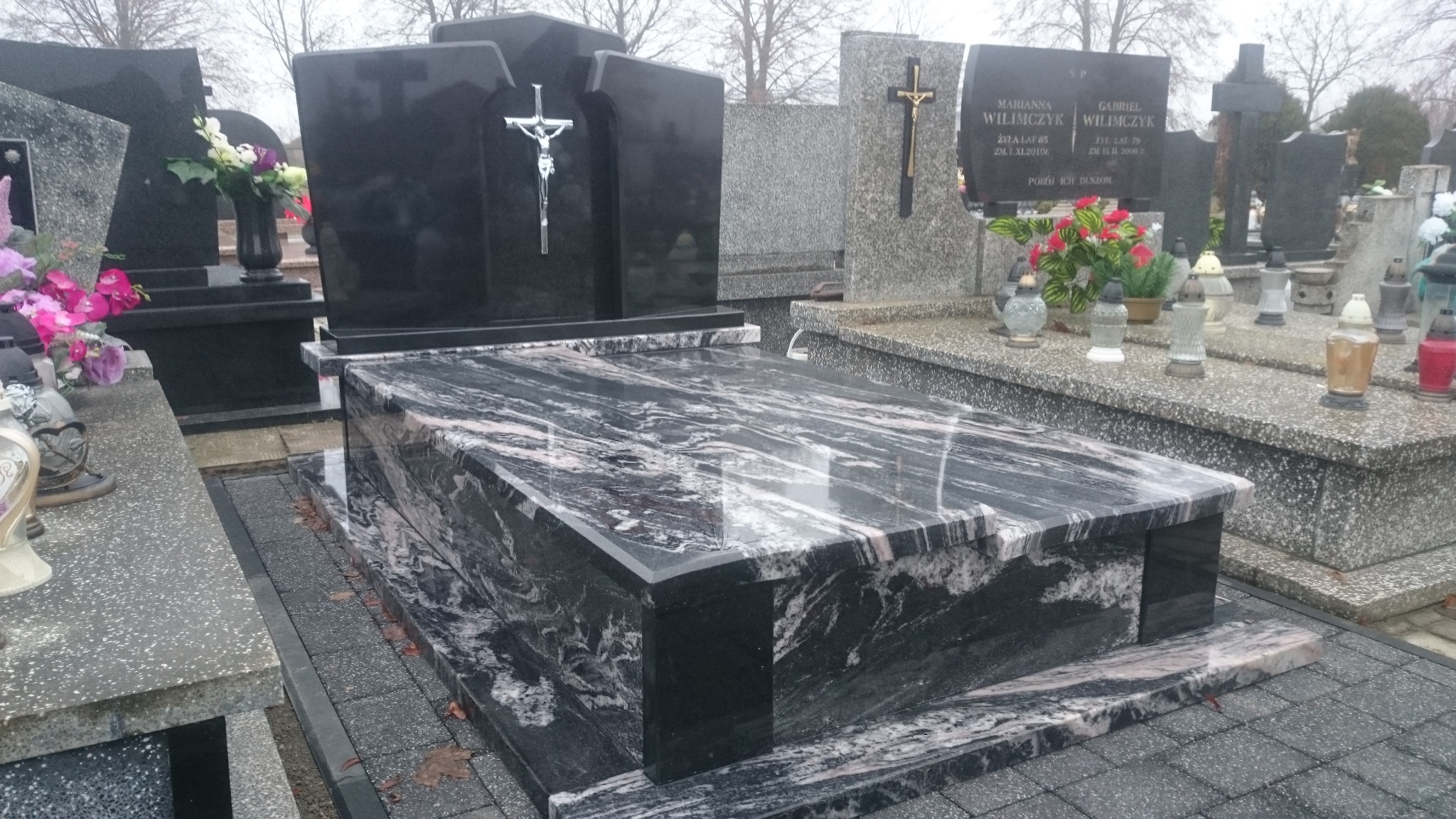 Czarny, marmurkowy pomnik z krzyżem na cmentarzu, widoczny w pochmurny dzień, z innymi nagrobkami w tle.