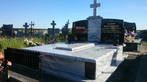 Jasny, marmurkowy pomnik z czarną tablicą inskrypcyjną i krzyżem, na tle cmentarza z licznymi nagrobkami i zielenią.