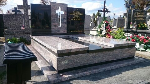 Nowoczesny, dwupoziomowy pomnik z jasnego granitu z czarnymi wstawkami, z wyciętym krzyżem w tle, na tle innych nagrobków na cmentarzu.