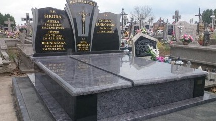 Granitowy pomnik nagrobny z inskrypcjami, krzyżem i napisem 'Jezu Ufam Tobie', na cmentarzu z licznymi innymi nagrobkami w tle.