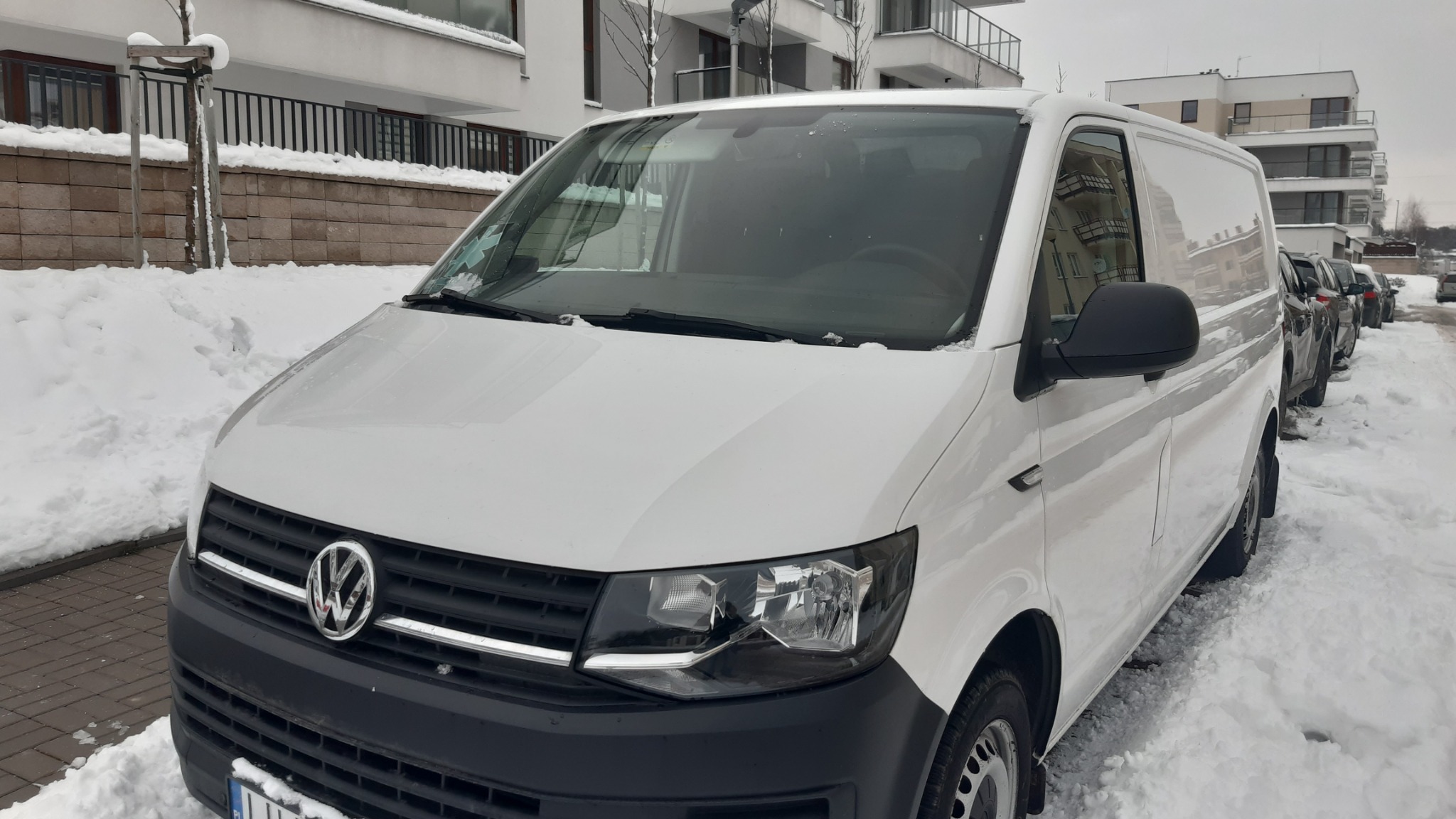 Biały samochód dostawczy Volkswagen zaparkowany na zasypanej śniegiem ulicy w pobliżu nowoczesnego budynku mieszkalnego.