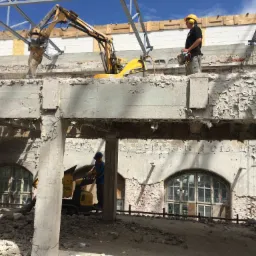 Dwóch robotników obsługuje maszyny do kruszenia betonu na terenie rozbieranego budynku z widocznymi łukowymi oknami i stertą gruzu.