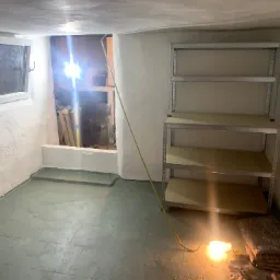 Piwnica po remoncie z pomalowanymi ścianami i podłogą, widoczne metalowe regały i prowizoryczne oświetlenie halogenowe.