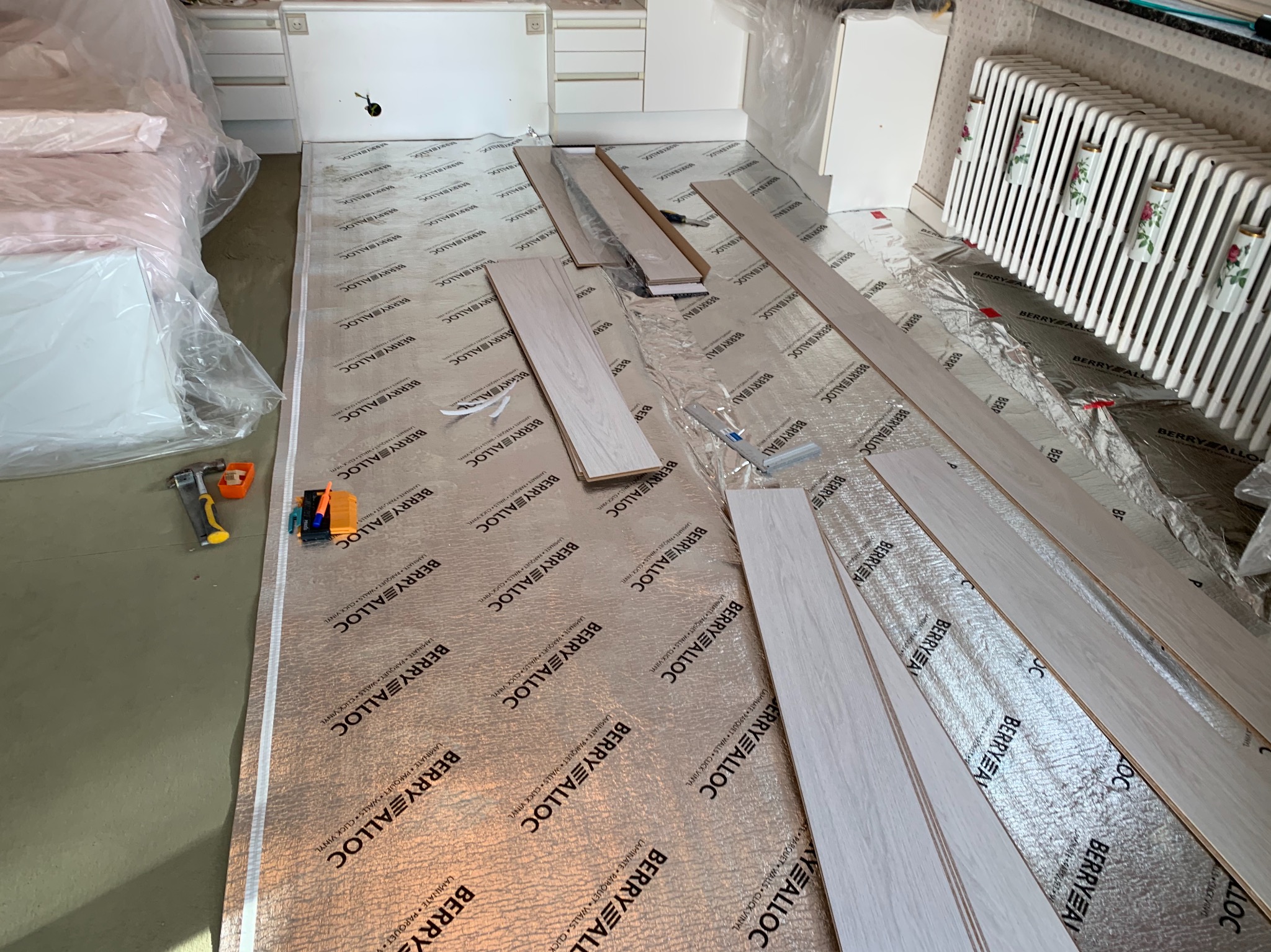 Przygotowanie do montażu paneli podłogowych: rozłożona folia izolacyjna z nadrukiem producenta i częściowo ułożone jasne panele, widoczne narzędzia montażowe.