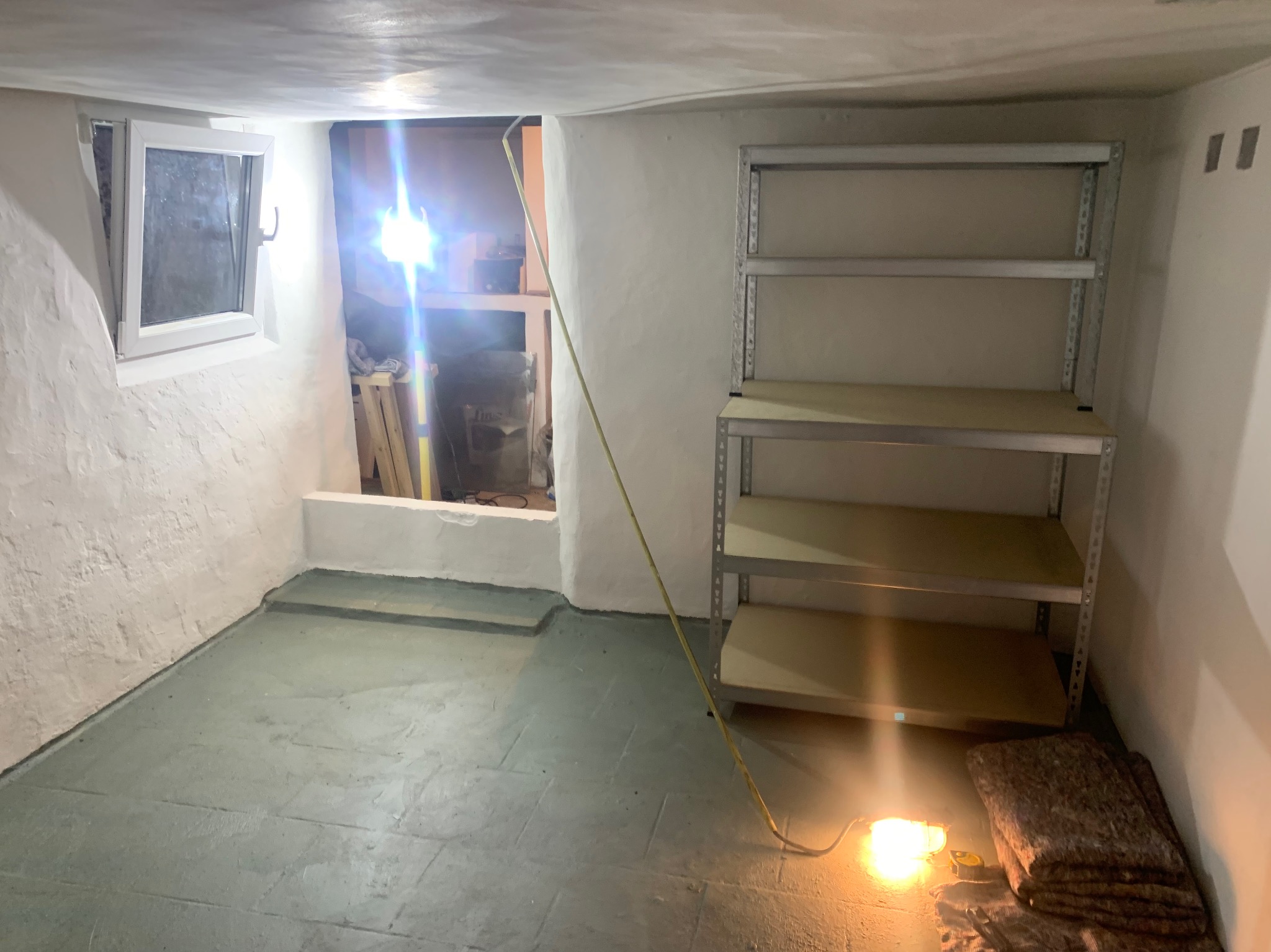 Piwnica po remoncie z pomalowanymi ścianami i podłogą, widoczne metalowe regały i prowizoryczne oświetlenie halogenowe.