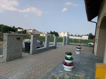 Ułożona kostka brukowa na podjeździe z elementami dekoracyjnymi w postaci donic z kwiatami i niskiego murku z kamienia łupanego, widok na dom i okoliczną zabudowę.