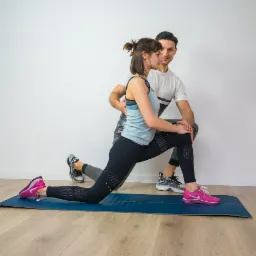 Kobieta w sportowym stroju wykonuje rozciąganie na macie, wspierana przez mężczyznę w białej koszulce.