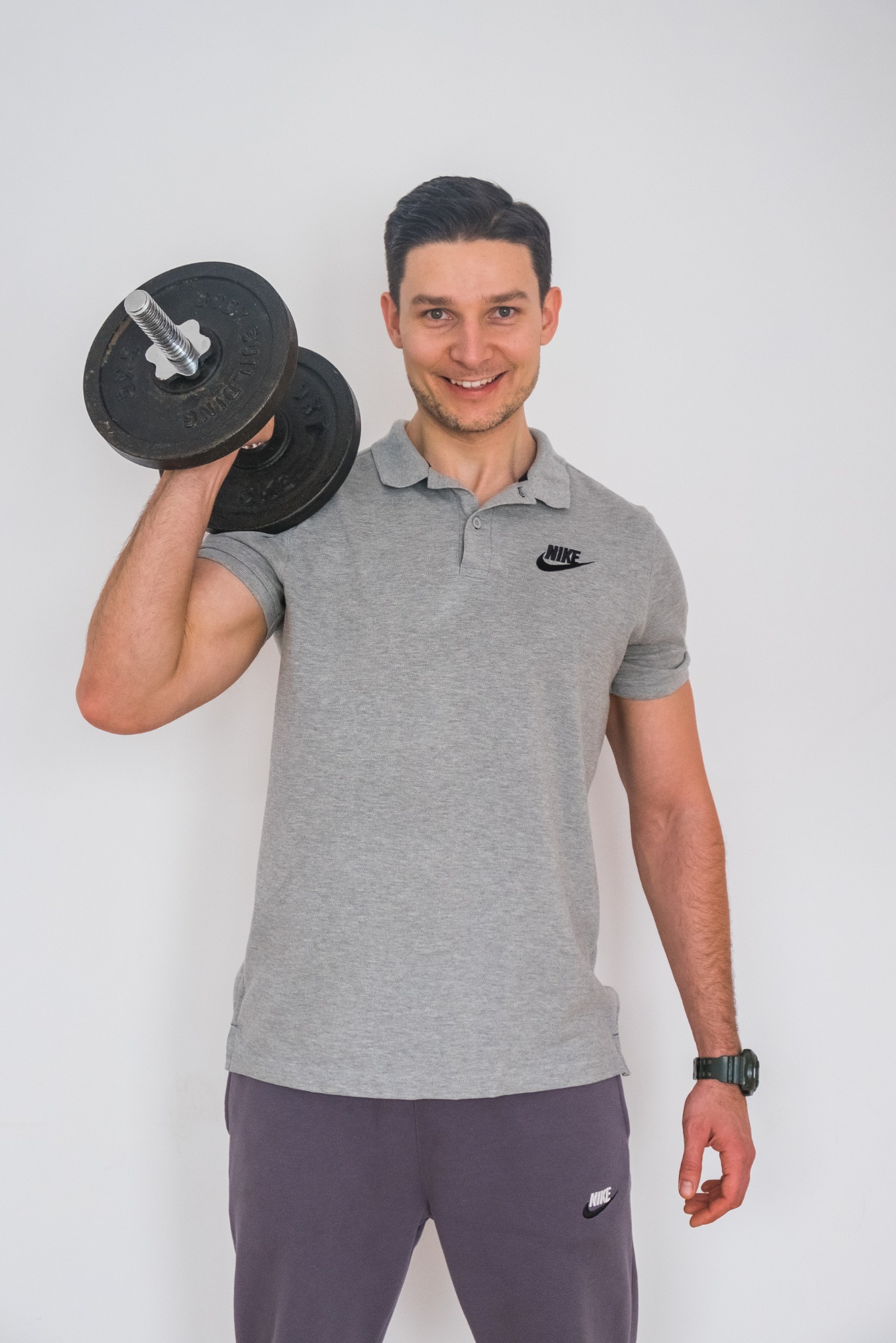 Uśmiechnięty mężczyzna w szarej koszulce polo z logo Nike, trzymający hantlę nad ramieniem na tle białej ściany.