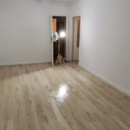 Wnętrze pokoju po remoncie z nową podłogą z paneli, widoczna lampa budowlana oświetlająca pomieszczenie i świeżo pomalowane białe ściany.