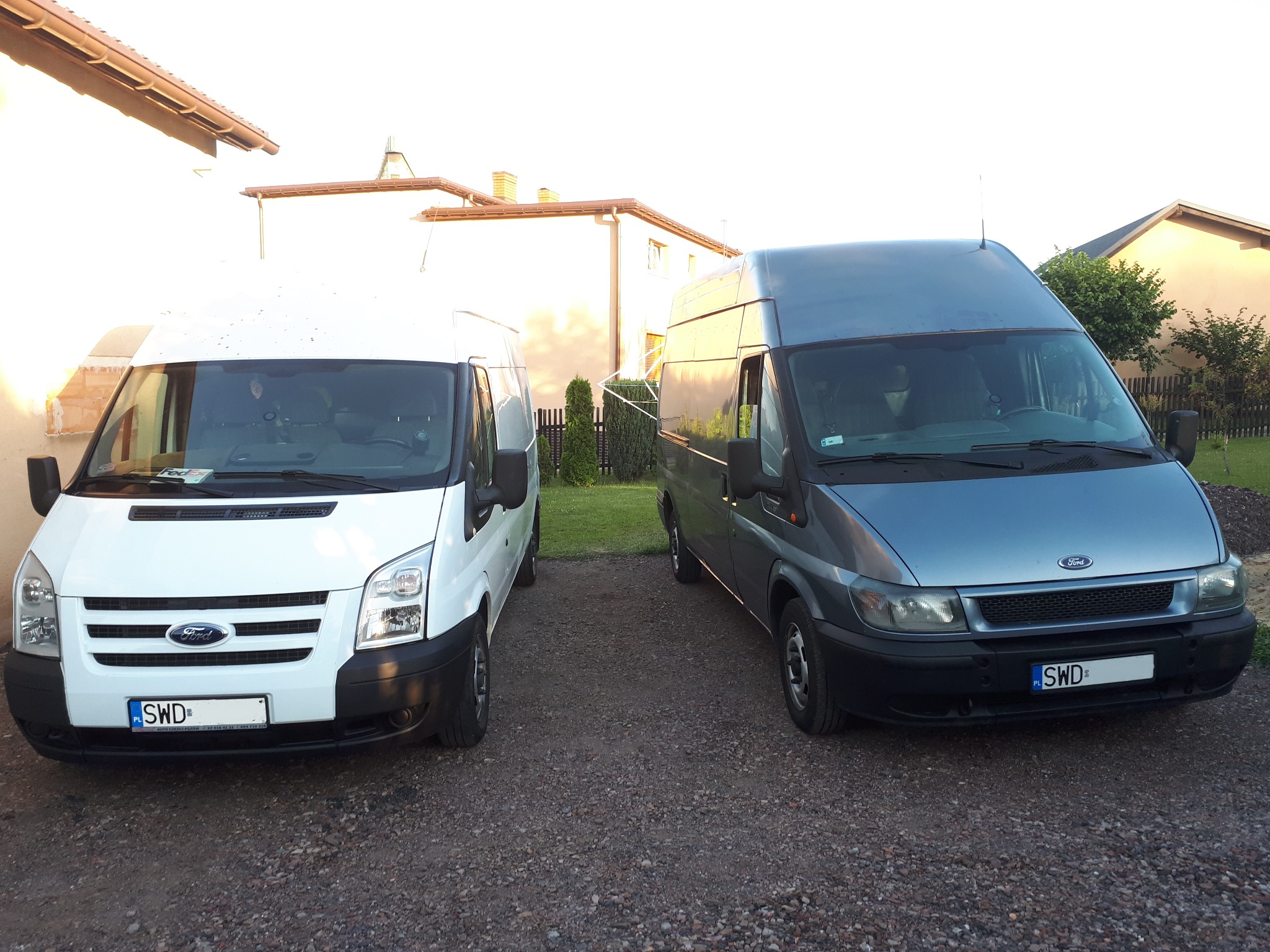 Dwa busy Ford Transit, biały i szary, zaparkowane na żwirowym podjeździe w Wodzisławiu Śląskim, z widocznymi tablicami rejestracyjnymi.