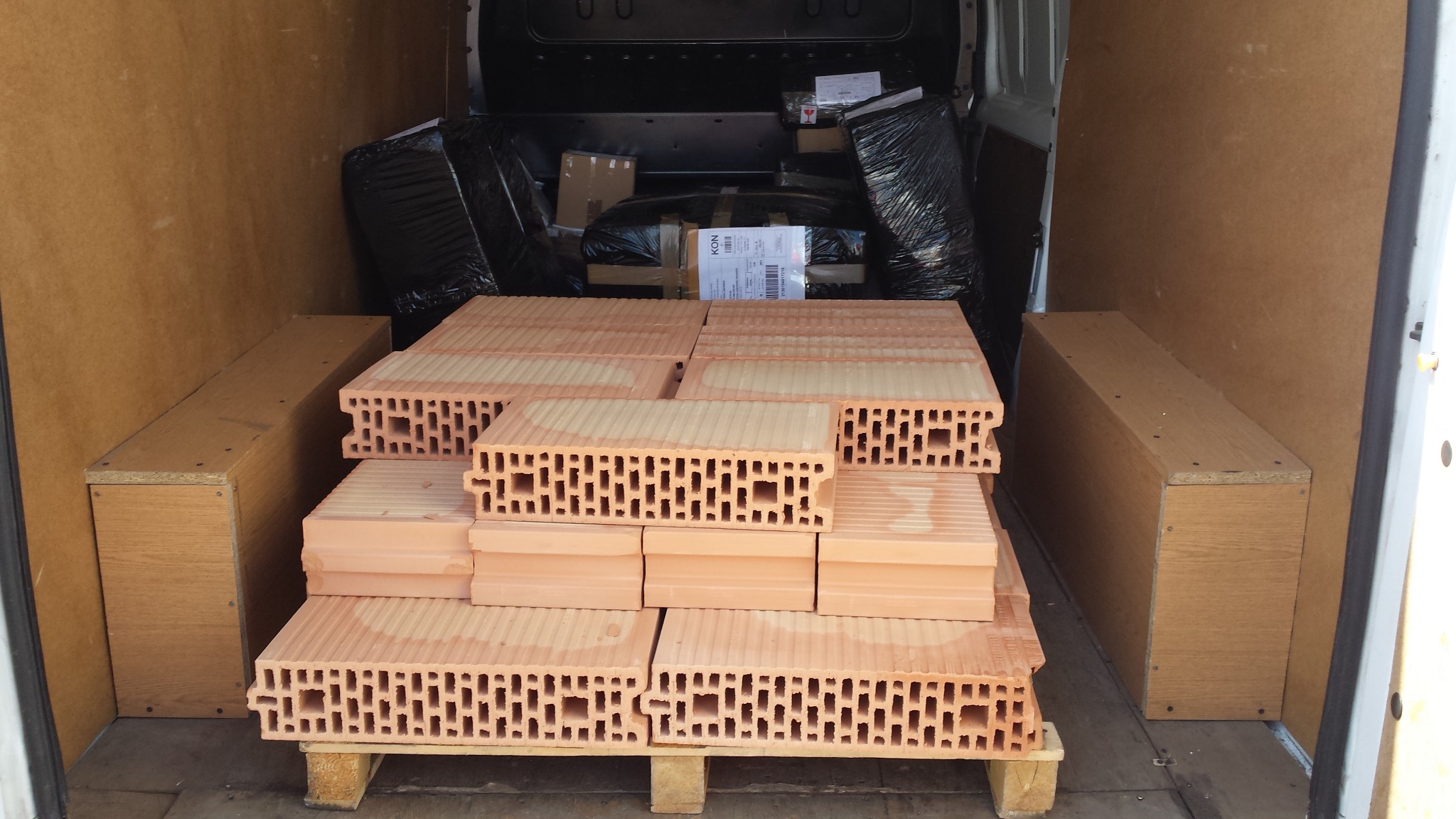 Cegły ceramiczne porotherm ułożone na palecie w przestrzeni ładunkowej busa, obok zapakowane paczki i meble, gotowe do transportu.