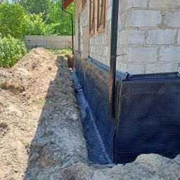 Fundament budynku w trakcie zabezpieczania: wykop obok ściany fundamentowej, czarna folia kubełkowa izolująca fundament, widoczny drenaż.