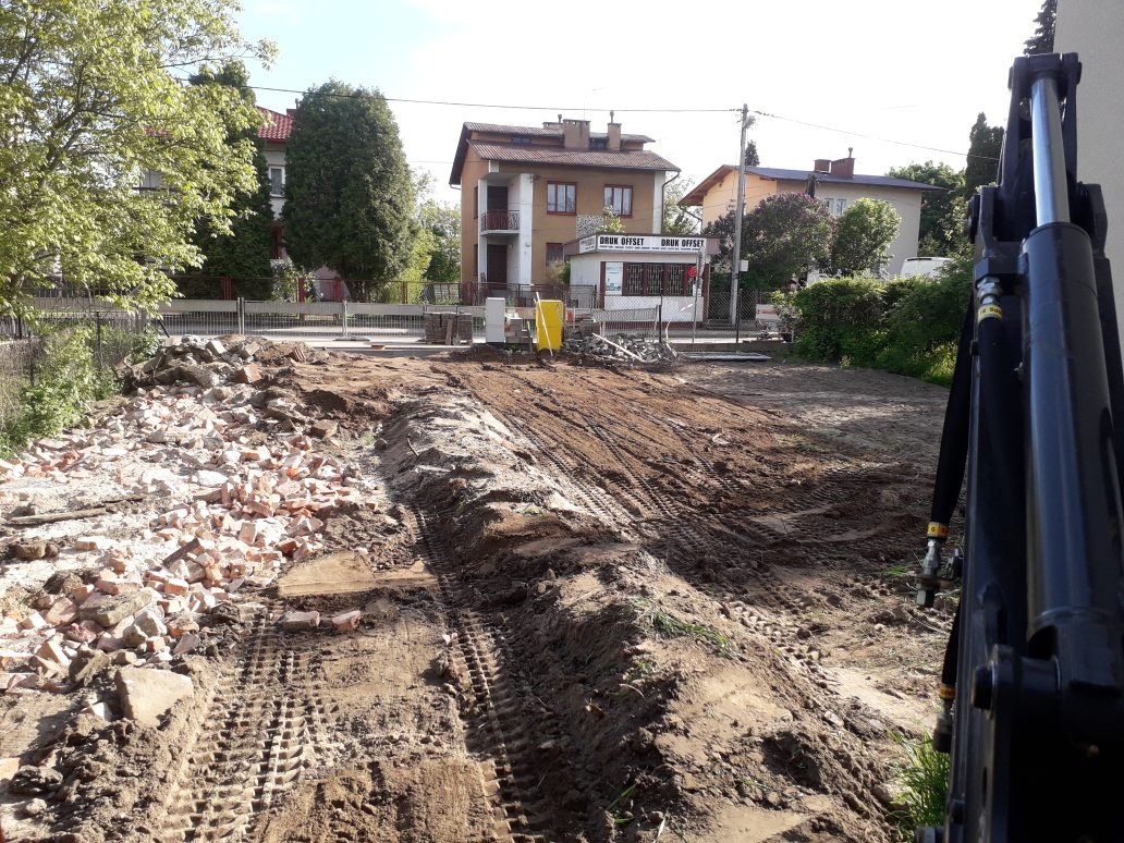Przygotowanie terenu pod budowę: wykarczowany teren z resztkami gruzu i śladami opon ciężkiego sprzętu, widoczna łyżka koparki w prawym dolnym rogu, w tle budynki mieszkalne.