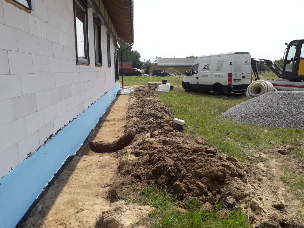 Fundament budynku z izolacją w kolorze niebieskim, wykopany wąski rów wzdłuż ściany, na trawie sterta żwiru i zaparkowany biały bus z logo firmy oraz mała koparka.
