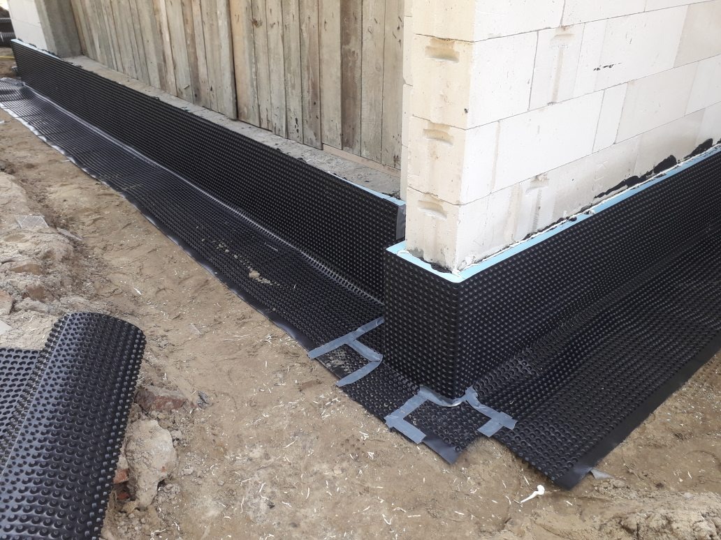 Czarna folia kubełkowa zabezpiecza fundament budynku z bloczków betonowych, widoczne łączenia taśmą, fragment zwiniętej rolki folii na ziemi.