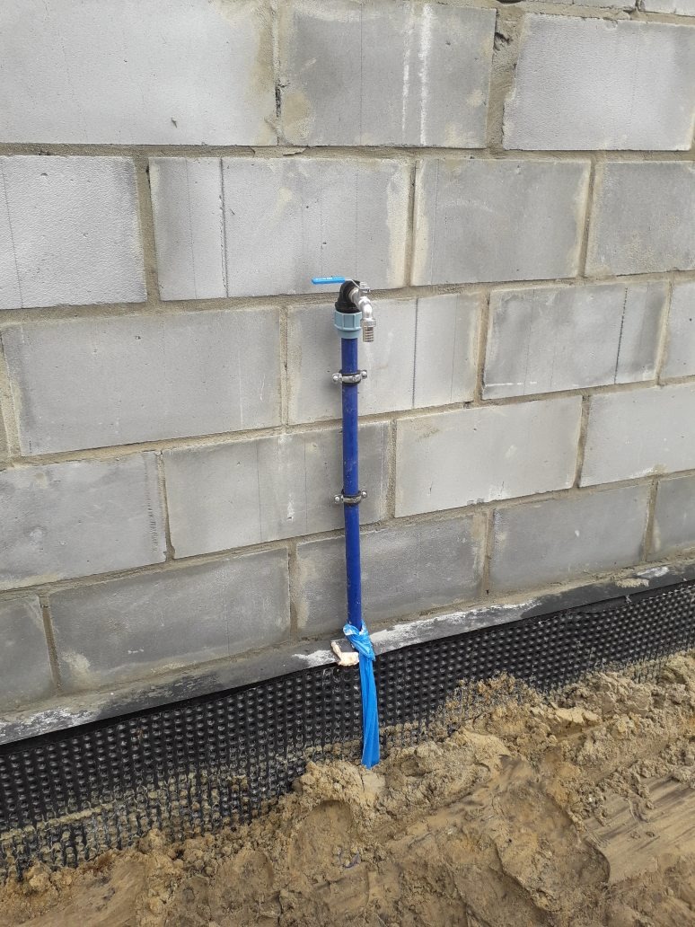 Niebieska rura z kranem wystająca z ziemi przy ścianie z bloczków betonowych i czarną folią izolacyjną fundamentu, zabezpieczona niebieską taśmą.