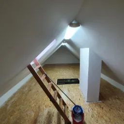 Poddasze ze skośnym sufitem po malowaniu, widoczne oświetlenie, schody drabinkowe i wyłaz dachowy.