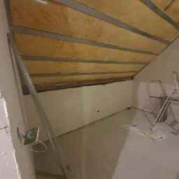 Poddasze w trakcie adaptacji: widoczne ocieplenie wełną mineralną pomiędzy metalowymi profilami, przygotowane pod montaż płyt gipsowo-kartonowych, surowa ściana z wystającą puszką elektryczną...