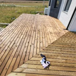 Nowo wybudowany taras z desek drewnianych, z widocznym fragmentem budynku w tle i pozostawioną parą skarpet na powierzchni.