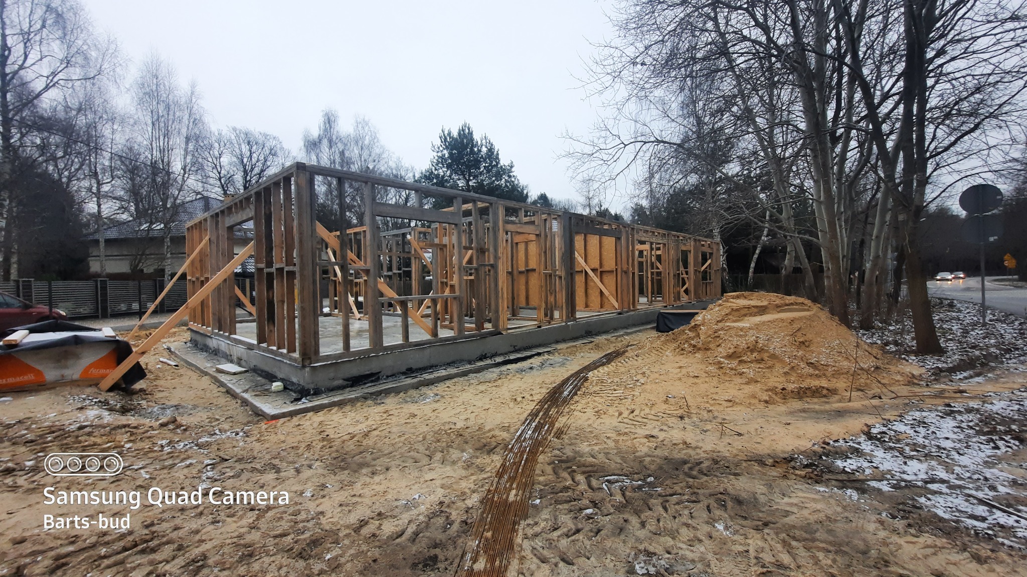 Surowy stan domu szkieletowego z drewnianą konstrukcją na betonowym fundamencie, widok na plac budowy z hałdą piasku i częściowo ośnieżonym terenem, drzewa w tle.