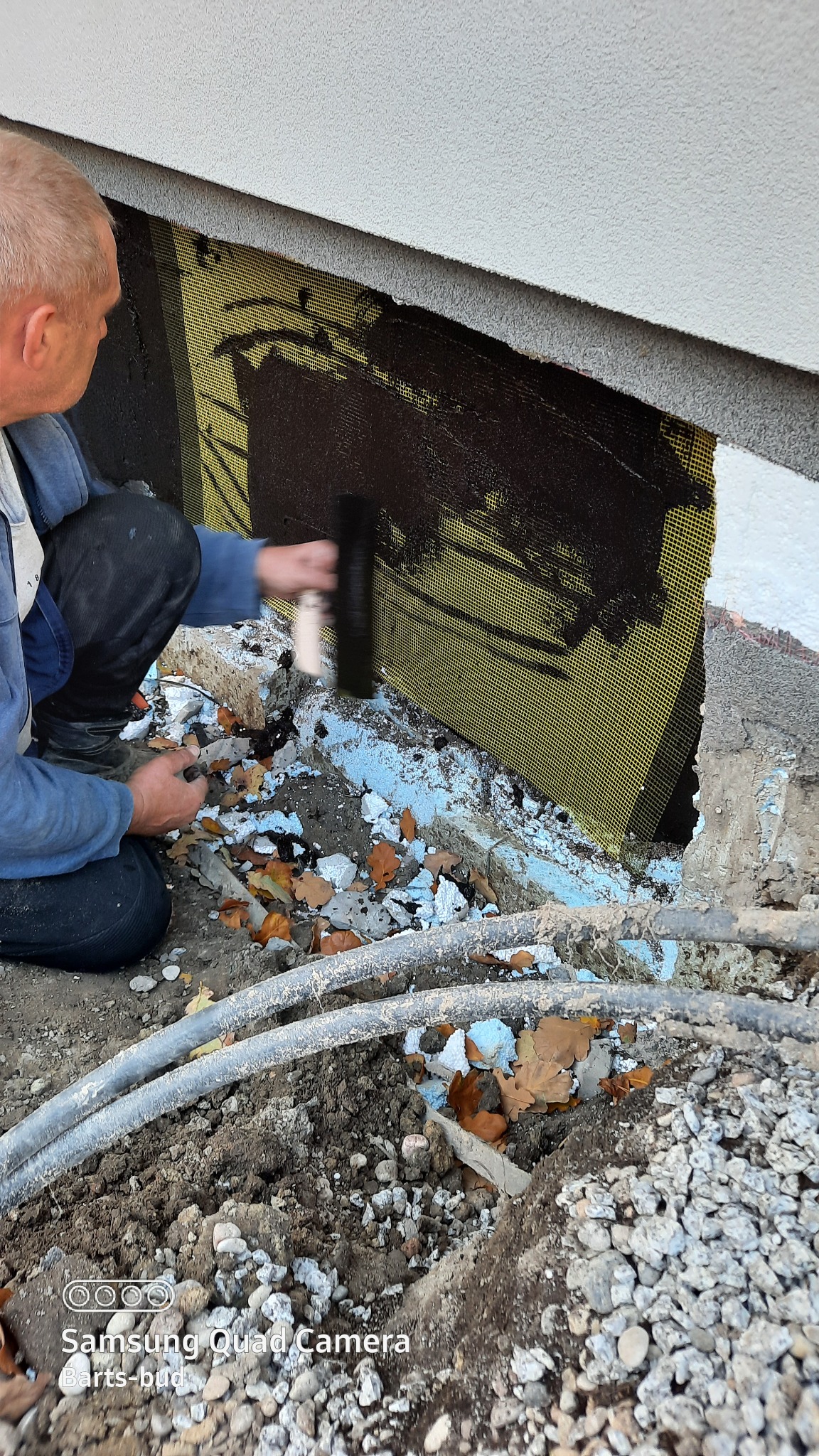 Prace przy izolacji pionowej fundamentu: aplikacja bitumicznej masy hydroizolacyjnej na siatkę wzmacniającą, widoczne fragmenty styropianu i rury w wykopie.