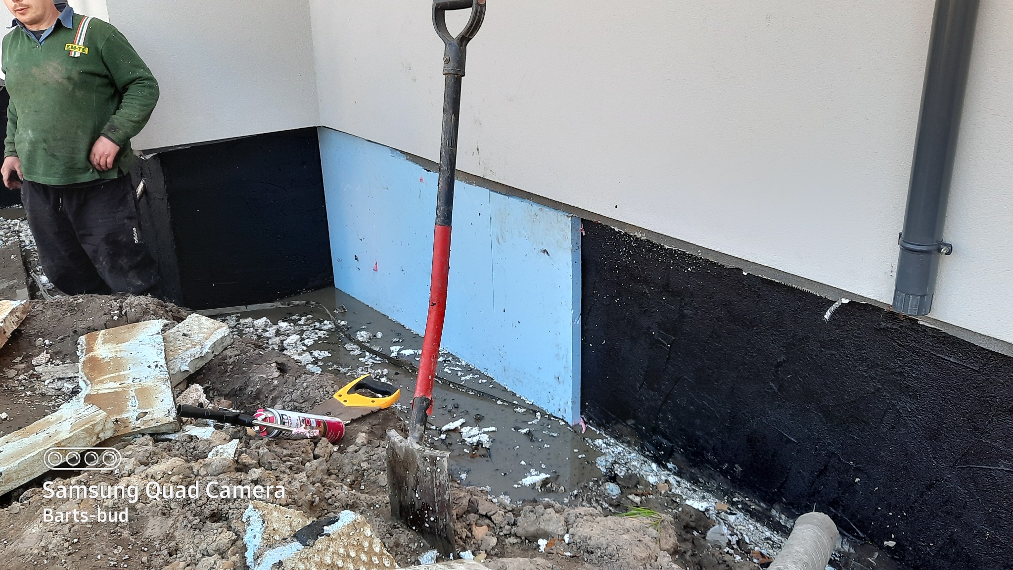 Prace izolacyjne fundamentów: zabezpieczenie ścian fundamentowych czarną masą bitumiczną i niebieskim styropianem, widoczna łopata, narzędzia i fragment pracownika.