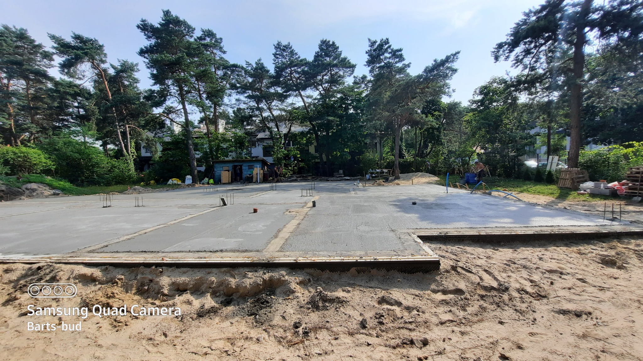 Wylany fundament z wystającymi prętami zbrojeniowymi na piaszczystym terenie, z widocznym szalunkiem i zielenią w tle.