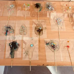 Ręcznie robione lizaki na patyku, każdy z unikalną dekoracją z posypek, lukru i ozdobnych elementów, ułożone na drewnianym blacie na przezroczystych arkuszach.