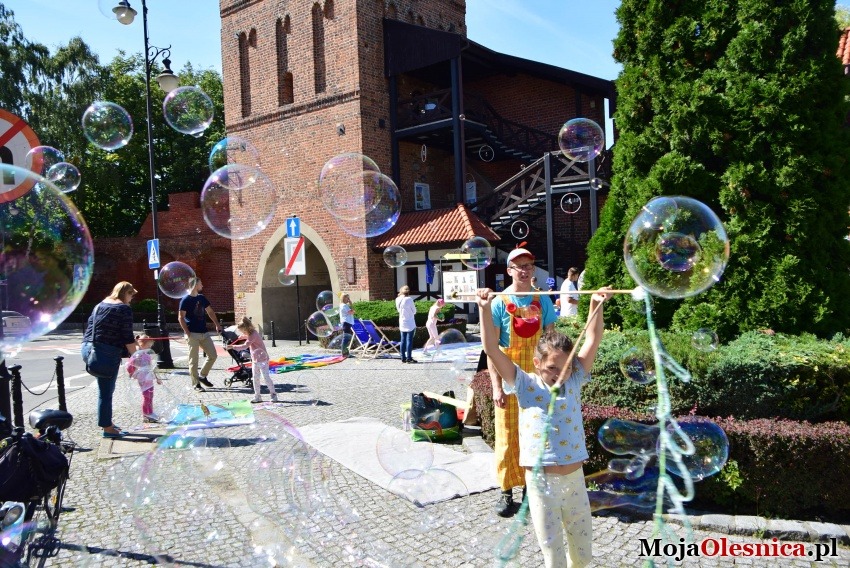 Dziewczynka w jasnych spodniach i koszulce robi duże bańki mydlane, obok klaun w żółto-czerwonym stroju, w tle ceglana wieża i bawiące się dzieci.