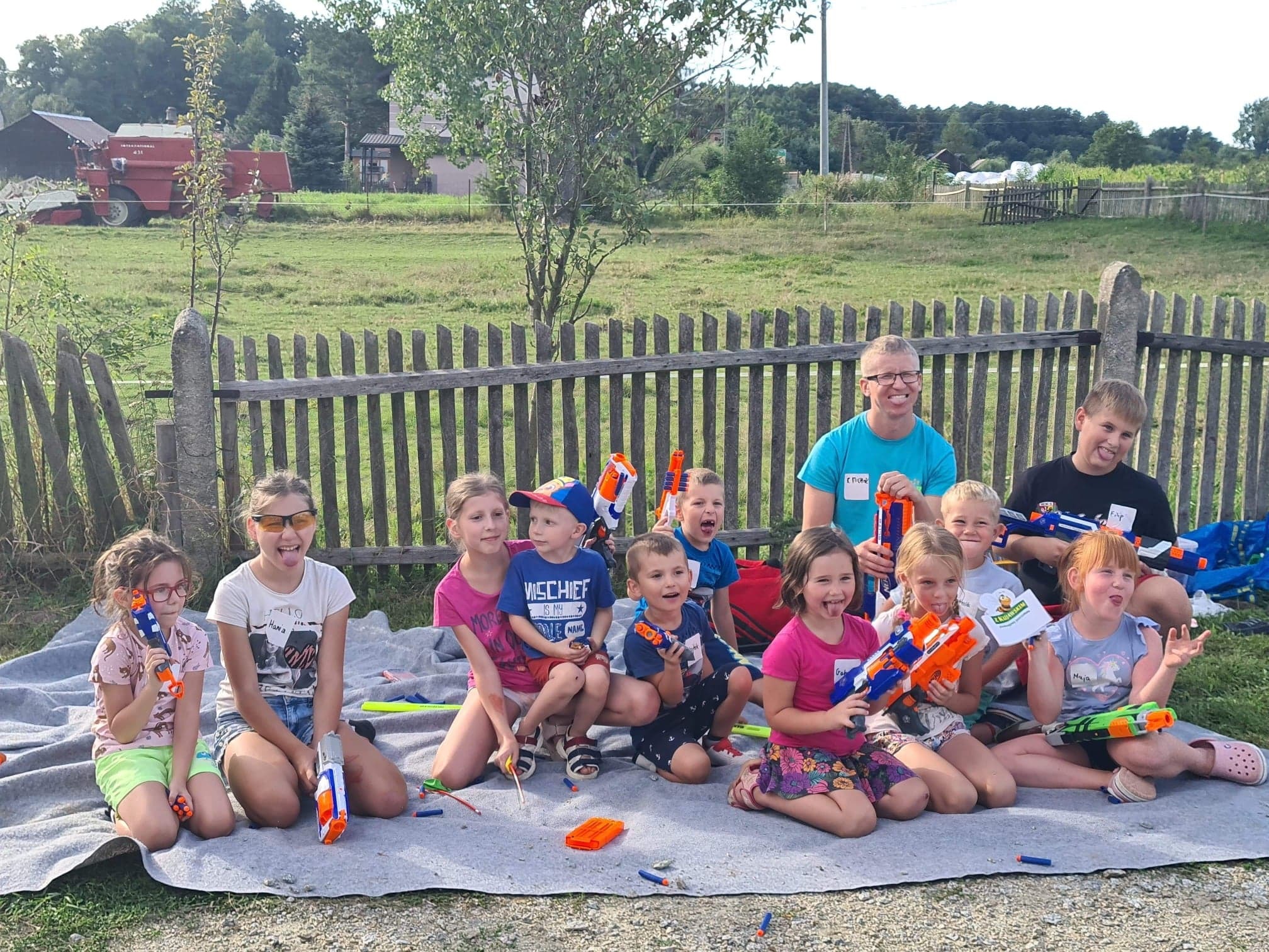 Grupa dzieci i dorosły siedzą na kocu na trawie, trzymając zabawki imitujące broń, w tle drewniany płot i pole z kombajnem.